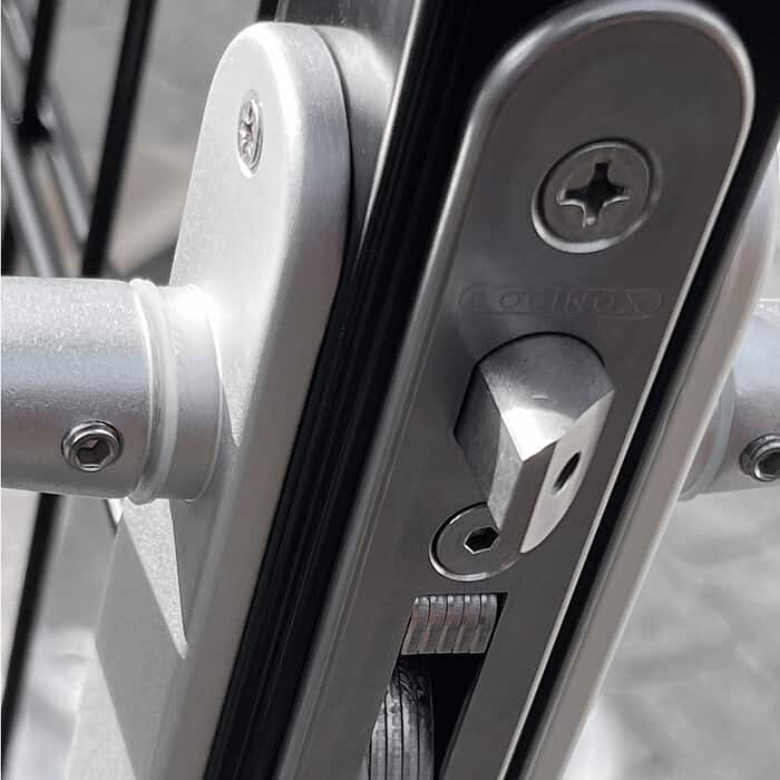 Nærbillede af en sølv dørlåsemekanisme med synlige skruer og en LOBP (Lift Off Bolt) på siden, monteret på en Metallåge 2D Panelfyld 6/5/6mm Sort Ral 9005 Komplet. Låsen er aktiveret og viser bolten udstrakt. Billedet har et gråtonefilter, der fremhæver de metalliske komponenter mod en let sløret baggrund.