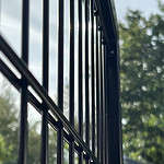 Nærbillede af den lodrette Metallåge 2D Panelfyld 6/5/6mm Sort RAL 9005 Komplet rækværk med en sløret baggrund af træer og himmel, der fanger lysreflektioner på metalstængerne.