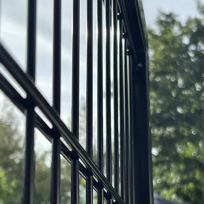 Nærbillede af den lodrette Metallåge 2D Panelfyld 6/5/6mm Sort RAL 9005 Komplet rækværk med en sløret baggrund af træer og himmel, der fanger lysreflektioner på metalstængerne.