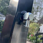 Nærbillede af en skinnende sekskantet bolt fastgjort til en sort metalstolpe, der matcher Metallåge 2D Panelfyld 6/5/6mm Sort Ral 9005 Komplet, med en udendørs haveindstilling sløret i baggrunden. Bolten er placeret perfekt ved hjørnet af denne robuste Metallåge.