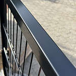 Nærbillede af en Metallåge 2D Panelfyld 6/5/6mm Sort Ral 9005 Komplet med lodrette stænger, der kaster skygger på jorden nedenunder. Jorden er belagt med rektangulære sten, og fokus er på rækværkets glatte, blanke overflade.