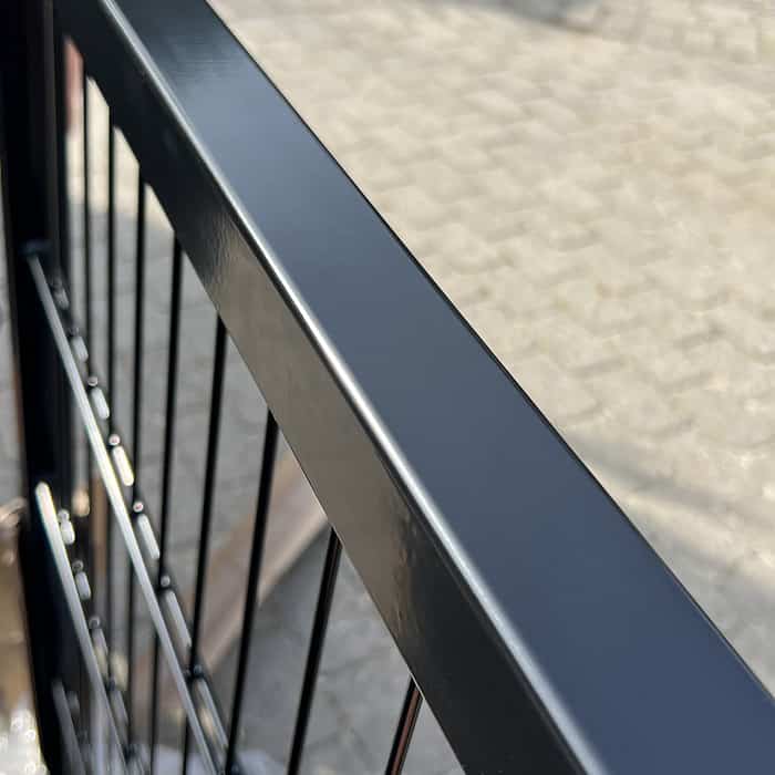 Nærbillede af en Metallåge 2D Panelfyld 6/5/6mm Sort Ral 9005 Komplet med lodrette stænger, der kaster skygger på jorden nedenunder. Jorden er belagt med rektangulære sten, og fokus er på rækværkets glatte, blanke overflade.