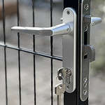 Nærbillede af Metallåge 2D Panelfyld 6/5/6mm Sort Ral 9005 Komplet portlås med nøgle indsat, drejet til venstre. Porten har lodrette og vandrette metalstænger, og baggrunden viser et ude af fokus brolagt område, hvilket tyder på en udendørs indstilling.