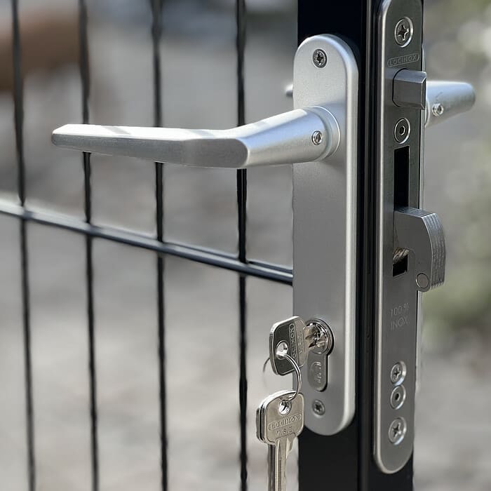 Nærbillede af Metallåge 2D Panelfyld 6/5/6mm Sort Ral 9005 Komplet portlås med nøgle indsat, drejet til venstre. Porten har lodrette og vandrette metalstænger, og baggrunden viser et ude af fokus brolagt område, hvilket tyder på en udendørs indstilling.