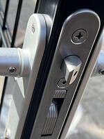 Et nærbillede af den sølvslåede låsemekanisme installeret på en Metallåge 2D Panelfyld 6/5/6mm Sort Ral 9005 Komplet sort metalport afslører dens indviklede detaljer. Låsen har en rektangulær bolt og en rund indsats til nøglen, med skruer og andre metalkomponenter tydeligt synlige, hvilket viser den detaljerede struktur mod overfladen af denne højkvalitetsport.