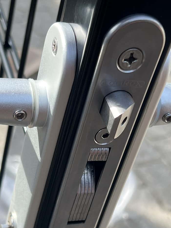 Et nærbillede af den sølvslåede låsemekanisme installeret på en Metallåge 2D Panelfyld 6/5/6mm Sort Ral 9005 Komplet sort metalport afslører dens indviklede detaljer. Låsen har en rektangulær bolt og en rund indsats til nøglen, med skruer og andre metalkomponenter tydeligt synlige, hvilket viser den detaljerede struktur mod overfladen af denne højkvalitetsport.