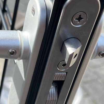 Et nærbillede af den sølvslåede låsemekanisme installeret på en Metallåge 2D Panelfyld 6/5/6mm Sort Ral 9005 Komplet sort metalport afslører dens indviklede detaljer. Låsen har en rektangulær bolt og en rund indsats til nøglen, med skruer og andre metalkomponenter tydeligt synlige, hvilket viser den detaljerede struktur mod overfladen af denne højkvalitetsport.