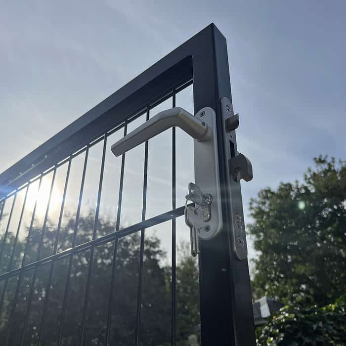 Et nærbillede af Metallåge 2D Panelfyld 6/5/6mm Sort Ral 9005 Komplet-port med låst håndtag mod en klar himmel. Sollys trænger gennem panelets stænger og kaster et subtilt skær rundt om porten. I baggrunden er træer og grønne områder oplyst af sollys.