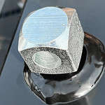 Nærbillede af en sekskantet metalmøtrik med et gevindhul, hvilende på en blank, reflekterende sort overflade, der ligner finishen på Metallåge 2D Panelfyld 6/5/6mm Sort RAL 9005 Komplet. Møtrikken har en sølvfarvet, let tekstureret finish og fremstår som ny og ubrugt.