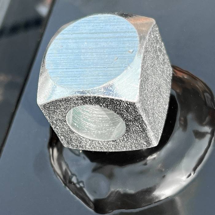 Nærbillede af en sekskantet metalmøtrik med et gevindhul, hvilende på en blank, reflekterende sort overflade, der ligner finishen på Metallåge 2D Panelfyld 6/5/6mm Sort RAL 9005 Komplet. Møtrikken har en sølvfarvet, let tekstureret finish og fremstår som ny og ubrugt.