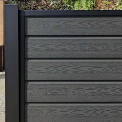 Nærbillede af en Komposit Hegn med sorte pulverlakeret stålstolper & u-skinner - Type 1 sammensat af mørke komposittræpaneler med vandrette planker, med metalramme. Grønt ser over hegnet og tilføjer dets moderne tiltrækningskraft.