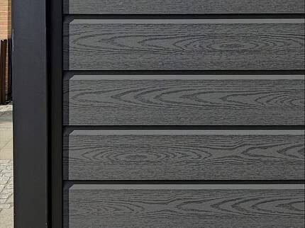 Nærbillede af en Komposit Hegn med sorte pulverlakeret stålstolper & u-skinner - Type 1 sammensat af mørke komposittræpaneler med vandrette planker, med metalramme. Grønt ser over hegnet og tilføjer dets moderne tiltrækningskraft.