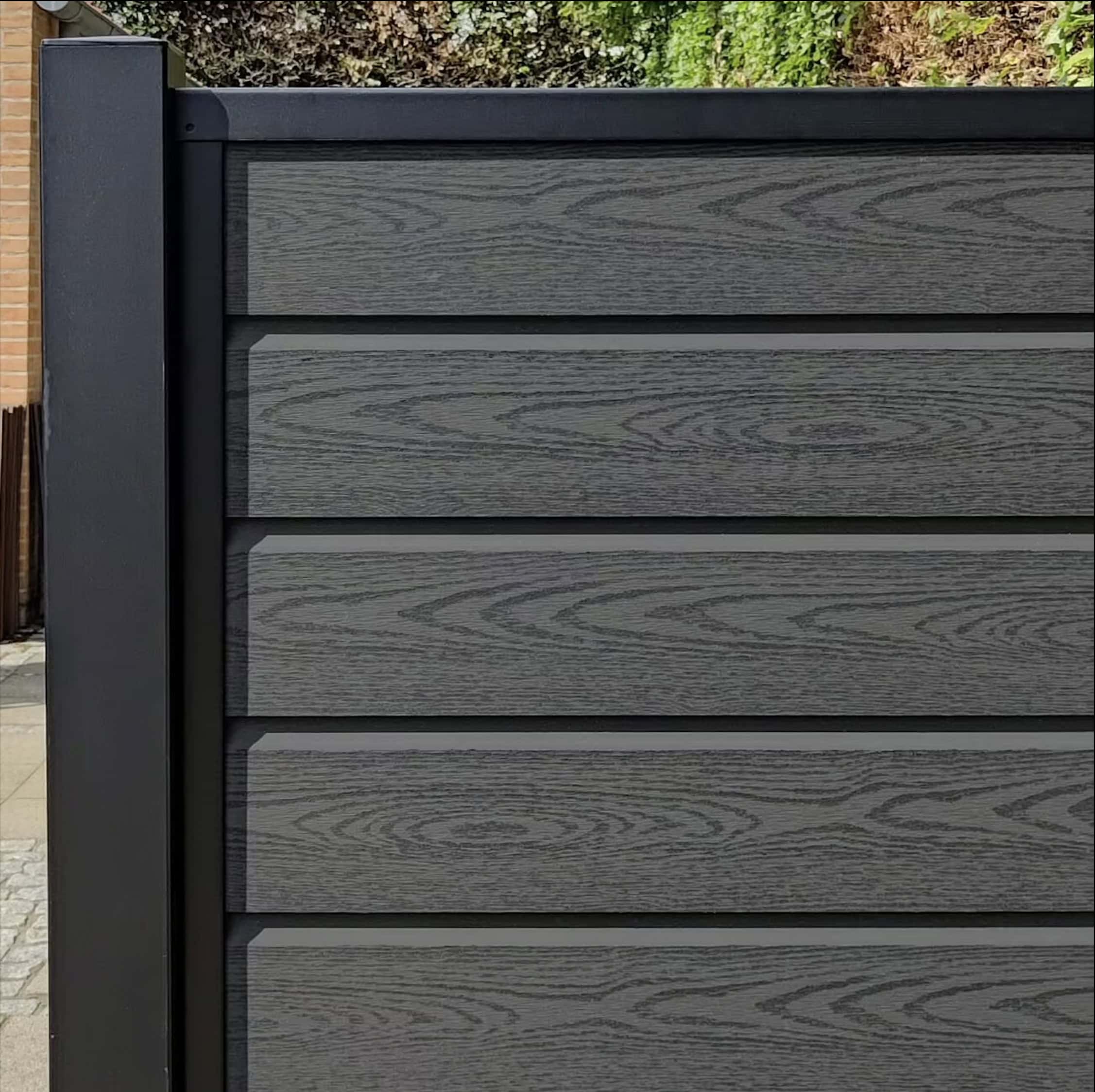 Nærbillede af en Komposit Hegn med sorte pulverlakeret stålstolper & u-skinner - Type 1 sammensat af mørke komposittræpaneler med vandrette planker, med metalramme. Grønt ser over hegnet og tilføjer dets moderne tiltrækningskraft.