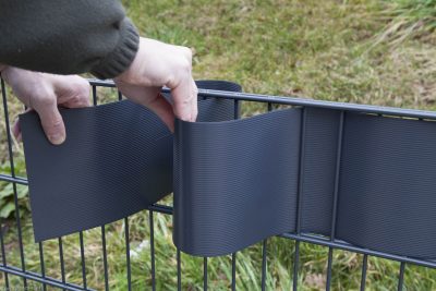 En person væver sorte plastikstrimler gennem stængerne på et metalhegn udendørs, hvilket skaber en Renovationsskjuler EcoHide Komplet. Personens hænder er synlige, der holder strimlerne, som bliver sat ind mellem de vandrette og lodrette bjælker. Græsset er synligt i baggrunden.