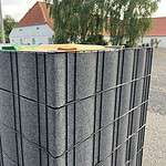 En stak mørkegrå paneler med teksturerede overflader er sikkert fastgjort med sorte metalrammer. Renovationsskjuler EcoHide Komplet er placeret udendørs på en grusoverflade, med bygninger med røde tegltage og et stort træ i baggrunden.