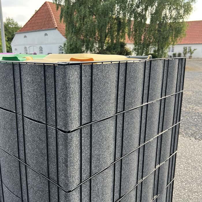 En stak mørkegrå paneler med teksturerede overflader er sikkert fastgjort med sorte metalrammer. Renovationsskjuler EcoHide Komplet er placeret udendørs på en grusoverflade, med bygninger med røde tegltage og et stort træ i baggrunden.