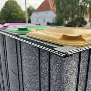 En række grå Renovationsskjuler EcoHide Komplet genbrugsspande med farvede låg, inklusive gul, grøn og lilla, er udstillet i en udendørs skraldespandsskjuler. Et hvidt hus med rødt tegltag og grønt er synligt i baggrunden på en delvist overskyet dag.