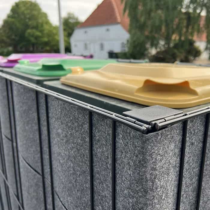 En række grå Renovationsskjuler EcoHide Komplet genbrugsspande med farvede låg, inklusive gul, grøn og lilla, er udstillet i en udendørs skraldespandsskjuler. Et hvidt hus med rødt tegltag og grønt er synligt i baggrunden på en delvist overskyet dag.