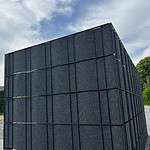 En Renovationsskjuler EcoHide Komplet, stor og mørkegrå med rektangulær form, med lodrette og vandrette paneler, står udendørs under en delvist overskyet himmel. Strukturen er beliggende på en grusoverflade, med træer og grønt synligt i baggrunden.