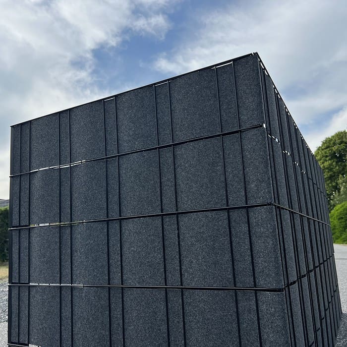 En Renovationsskjuler EcoHide Komplet, stor og mørkegrå med rektangulær form, med lodrette og vandrette paneler, står udendørs under en delvist overskyet himmel. Strukturen er beliggende på en grusoverflade, med træer og grønt synligt i baggrunden.