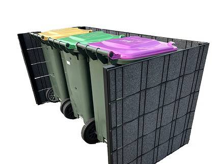 Tre grønne skraldespande på hjul med farvede låg (gul, grøn og lilla) er placeret i en rektangulær, mørkfarvet, paneleret Renovationsskjuler EcoHide Komplet. Indhegningen omgiver skraldespandene på tre sider, hvilket skaber et pænt og organiseret affaldsplads.