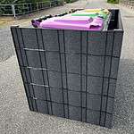 Et sidebillede af tre store skraldespande med låg i forskellige farver (lilla, gul og grøn) opbevaret i en sort Renovationsskjuler EcoHide Komplet på en asfalt overflade. Et metalhegn og grønt er synligt i baggrunden.