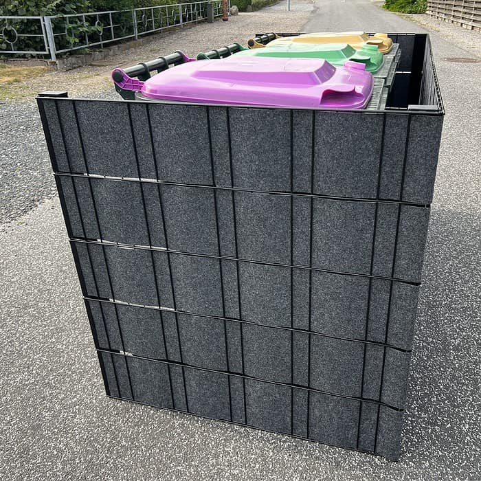 Et sidebillede af tre store skraldespande med låg i forskellige farver (lilla, gul og grøn) opbevaret i en sort Renovationsskjuler EcoHide Komplet på en asfalt overflade. Et metalhegn og grønt er synligt i baggrunden.