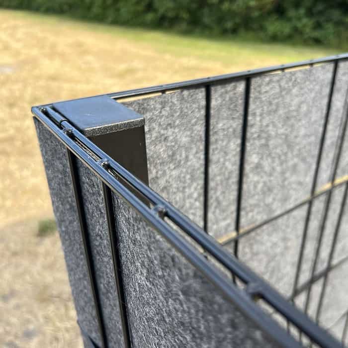 Et nærbillede af et trådhegn med sort hjørnestolpe, delvist dækket af grå stofpaneler. I det fjerne skjuler Renovationsskjuler EcoHide Komplet kunstfærdigt skraldespande mod en baggrund af tørt græs og træer.