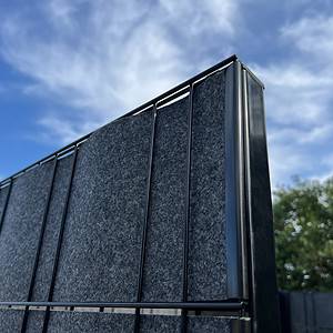 Nærbillede af det øverste hjørne af en Renovationsskjuler EcoHide Komplet, med sort metal foret med gråt stof, sat mod en blå himmel oversået med spredte hvide skyer. Baggrunden viser spidserne af grønne træer, hvilket gør den ideel til et Skraldespandsskjuler DIY-projekt.