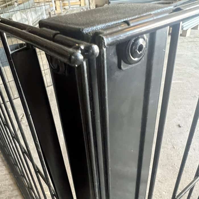 Nærbillede af et sort metalporthængsel med en låsemekanisme. Lågen og hængslet ser ud til at være en del af en robust, svejset struktur med synlige bolte. Baggrunden er en industriel indstilling med et betongulv og metalstænger, perfekt til at bruge Renovationsskjuler EcoHide Komplet i dit skraldespandsskjuler DIY-projekt.