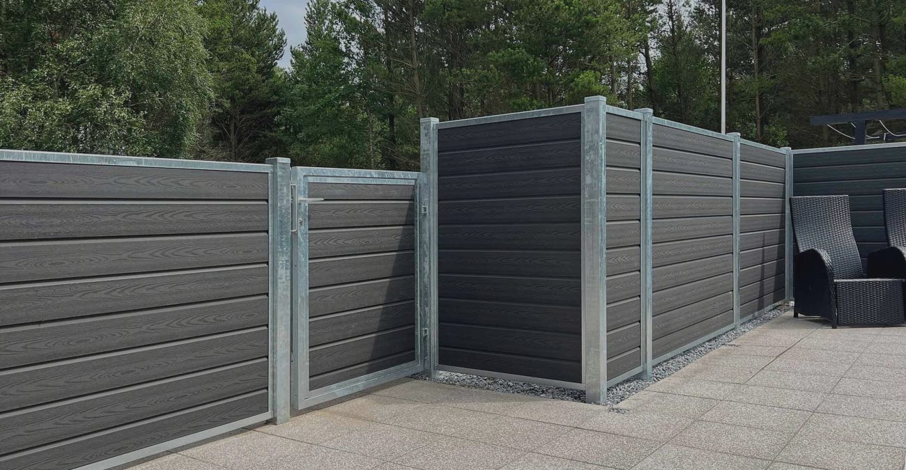 Et moderne udendørsområde kan prale af en grå komposithegn med vandrette lameller og metalliske stolper, der omslutter en terrasse med slanke grå fliser. Ud over hegnet strækker træer sig op under en overskyet himmel og skaber en elegant kontrast i denne private oase.