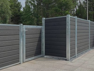 Et moderne udendørsområde kan prale af en grå komposithegn med vandrette lameller og metalliske stolper, der omslutter en terrasse med slanke grå fliser. Ud over hegnet strækker træer sig op under en overskyet himmel og skaber en elegant kontrast i denne private oase.