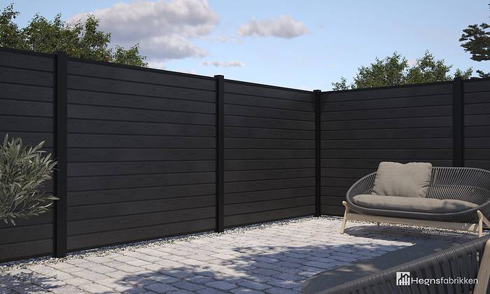 En moderne udendørs terrasse med et sort komposithegn med pulverlakerede stålstolper og U-spor (Type 1), grå polstrede siddepladser og et asfalteret stengulv. Himlen er delvist overskyet, med grønt fra træer synligt i baggrunden. I nederste højre hjørne fremhæver "Hegnsfabrikken"-logoet deres seneste Komposit Hegn-kollektionstilbud.