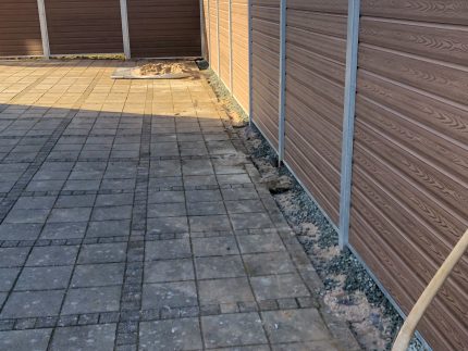 En brolagt gårdhave omgivet af et Komposit Hegn med sorte pulverlakeret stålstolper & u-skinner - Type 1. En spade ligger i jorden ved siden af hegnet og antyder landskabsarbejde. Træer og bygninger dukker op i baggrunden.