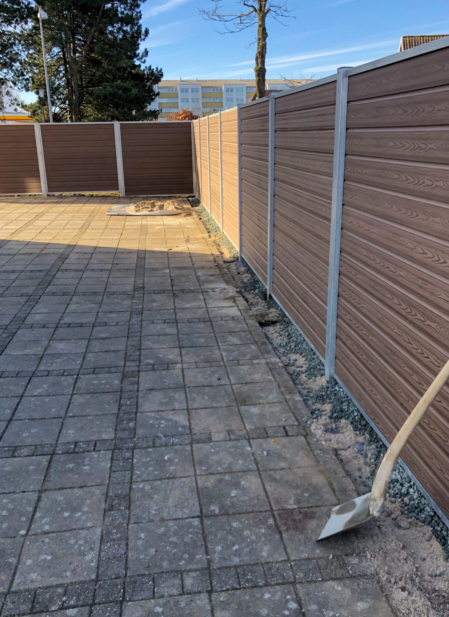 En brolagt gårdhave omgivet af et Komposit Hegn med sorte pulverlakeret stålstolper & u-skinner - Type 1. En spade ligger i jorden ved siden af hegnet og antyder landskabsarbejde. Træer og bygninger dukker op i baggrunden.