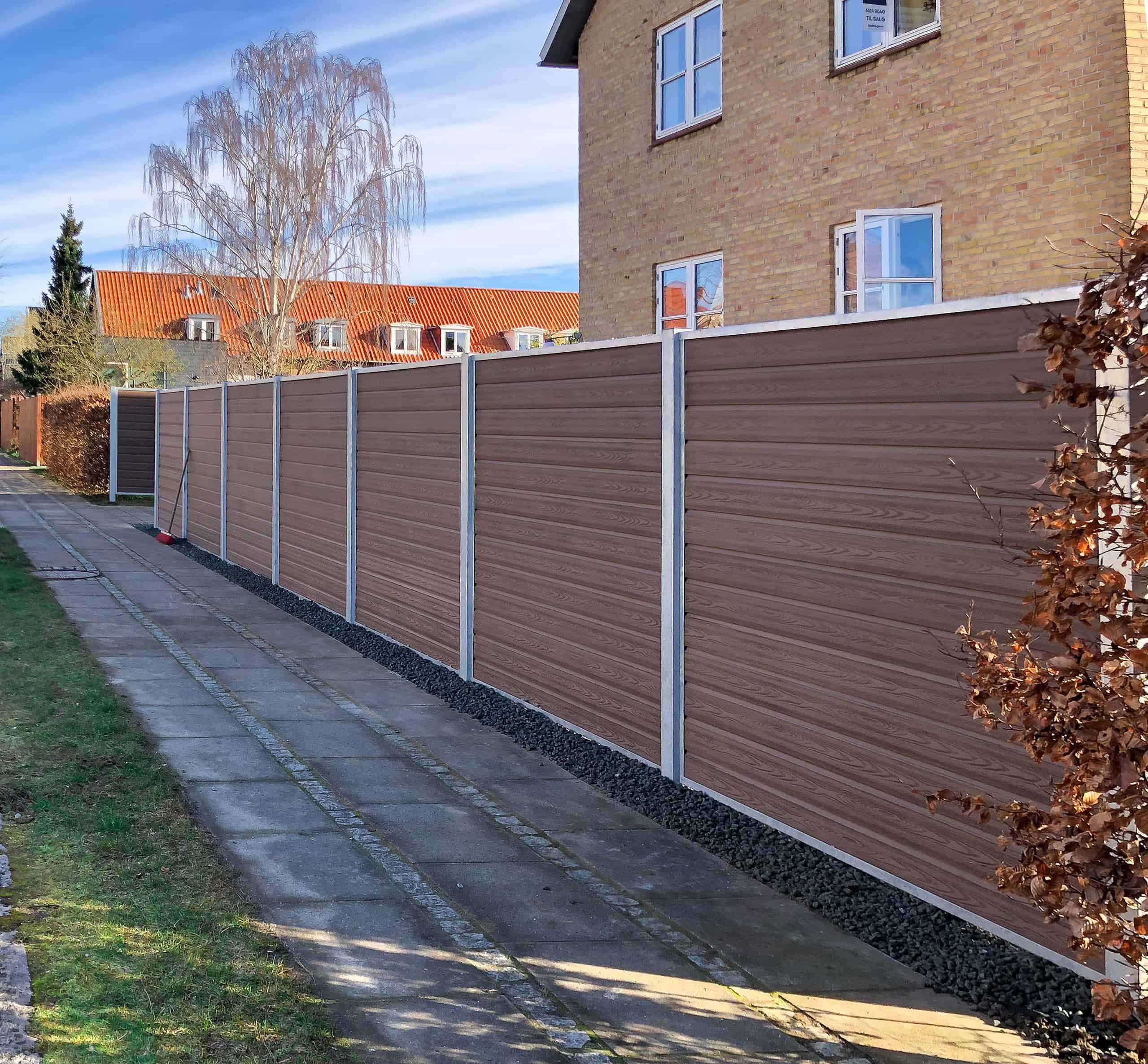 Komposit Hegn brun med stålstolper & u-skinner - Type 1 ligger langs en brolagt sti ved siden af et beige murstenshus med røde tagsten på en solrig dag. Sparsomme træer og buske dukker op langs stien.