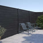 Moderne udendørs terrasse med to slanke metalstole med armlæn, placeret mod et højt, mørkt træpanelhegn. Området omfatter en potteplante til venstre og lidt grønt til højre under en klar blå himmel. Oplev vores Komposit Hegn med sorte pulverlakeret stålstolper & u-skinner - Type 1 for et stilfuldt touch til din udendørs.