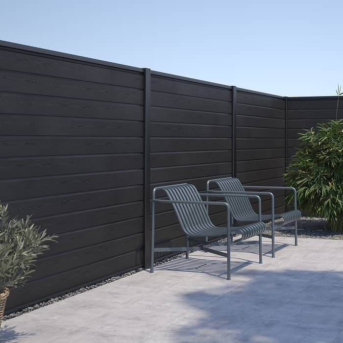 Moderne udendørs terrasse med to slanke metalstole med armlæn, placeret mod et højt, mørkt træpanelhegn. Området omfatter en potteplante til venstre og lidt grønt til højre under en klar blå himmel. Oplev vores Komposit Hegn med sorte pulverlakeret stålstolper & u-skinner - Type 1 for et stilfuldt touch til din udendørs.