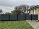 Komposit Hegn med sorte pulverlakerede stålstolper og skinner, bestående af mørke vandrette paneler og en port, står ved siden af en beige bygning. Det komplementerer den græsklædte jord og den lille stengårdhave, mens træer dukker op i baggrunden under en overskyet himmel.