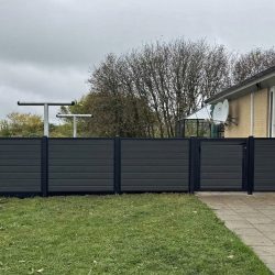 Komposit Hegn med sorte pulverlakerede stålstolper og skinner, bestående af mørke vandrette paneler og en port, står ved siden af en beige bygning. Det komplementerer den græsklædte jord og den lille stengårdhave, mens træer dukker op i baggrunden under en overskyet himmel.
