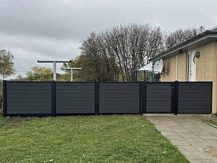 Komposit Hegn med sorte pulverlakerede stålstolper og skinner, bestående af mørke vandrette paneler og en port, står ved siden af en beige bygning. Det komplementerer den græsklædte jord og den lille stengårdhave, mens træer dukker op i baggrunden under en overskyet himmel.