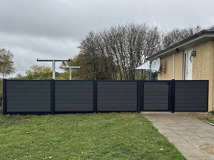 Komposit Hegn med sorte pulverlakerede stålstolper og skinner, bestående af mørke vandrette paneler og en port, står ved siden af en beige bygning. Det komplementerer den græsklædte jord og den lille stengårdhave, mens træer dukker op i baggrunden under en overskyet himmel.