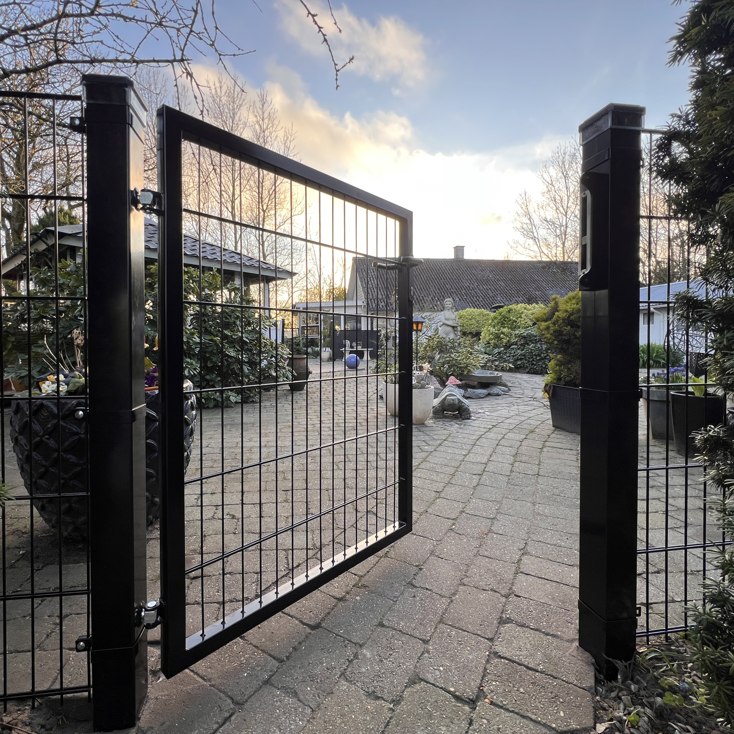 En Metallåge 2D Panelfyld 6/5/6mm Sort Ral 9005 Komplet står åben på den brolagte sti, der fører ind i en frodig have med planter og træer. Himlen er delvist skyet, og et hus ses i baggrunden.