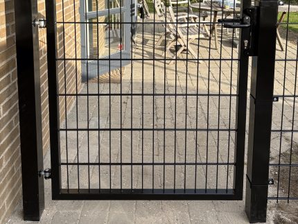 Metallåge 2D Panelfyld 6/5/6mm Sort Ral 9005 Komplet er monteret på to solide stolper og åbner op til en brolagt terrasse med havemøbler og frodig beplantning i baggrunden.