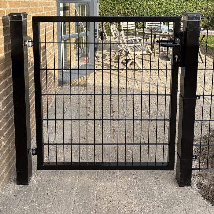 Metallåge 2D Panelfyld 6/5/6mm Sort Ral 9005 Komplet er monteret på to solide stolper og åbner op til en brolagt terrasse med havemøbler og frodig beplantning i baggrunden.