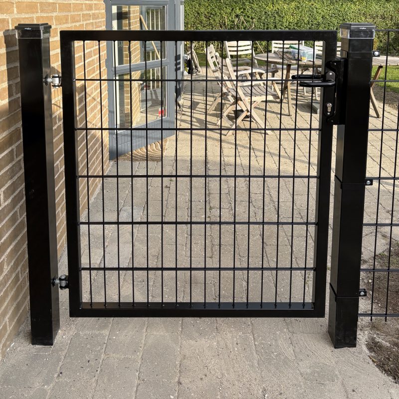 Metallåge 2D Panelfyld 6/5/6mm Sort Ral 9005 Komplet er monteret på to solide stolper og åbner op til en brolagt terrasse med havemøbler og frodig beplantning i baggrunden.