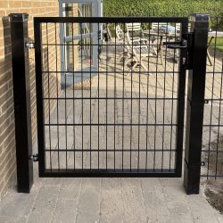Metallåge 2D Panelfyld 6/5/6mm Sort Ral 9005 Komplet er monteret på to solide stolper og åbner op til en brolagt terrasse med havemøbler og frodig beplantning i baggrunden.