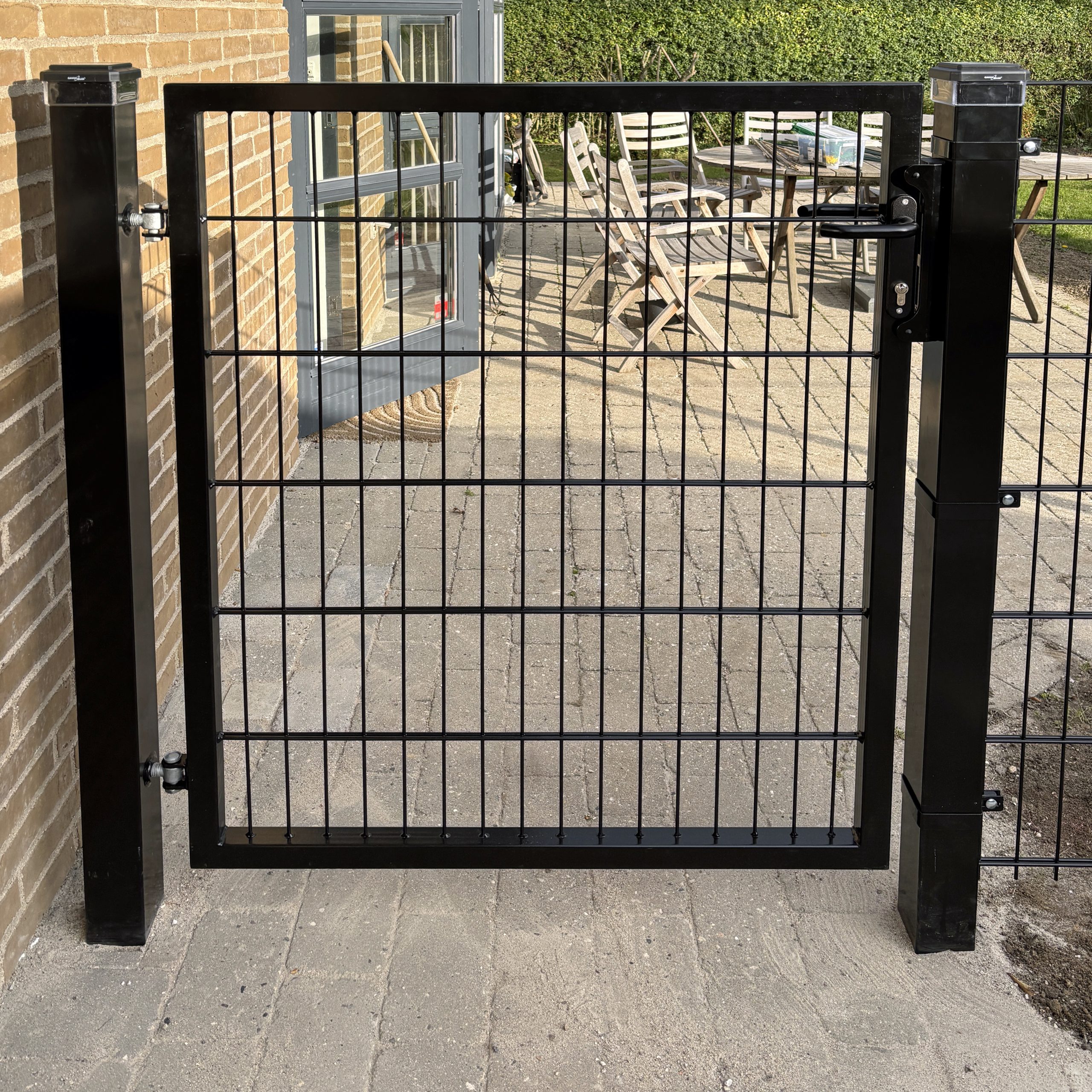 Metallåge 2D Panelfyld 6/5/6mm Sort Ral 9005 Komplet er monteret på to solide stolper og åbner op til en brolagt terrasse med havemøbler og frodig beplantning i baggrunden.