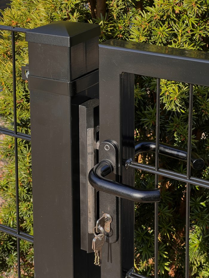 Et nærbillede af Metallåge 2D Panelfyld 6/5/6mm Sort Ral 9005 Komplet med lås og håndtag. Tre nøgler er sat i låsen, med synligt grønt løv i baggrunden.