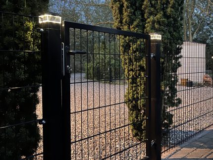 Et metallåge 2D panelfelt 6/5/6 mm sort Ral 9005 komplet med solcellelamper oven på stolperne, placeret blandt høje stedsegrønne træer og en grusjord, fotograferet i skumringen.